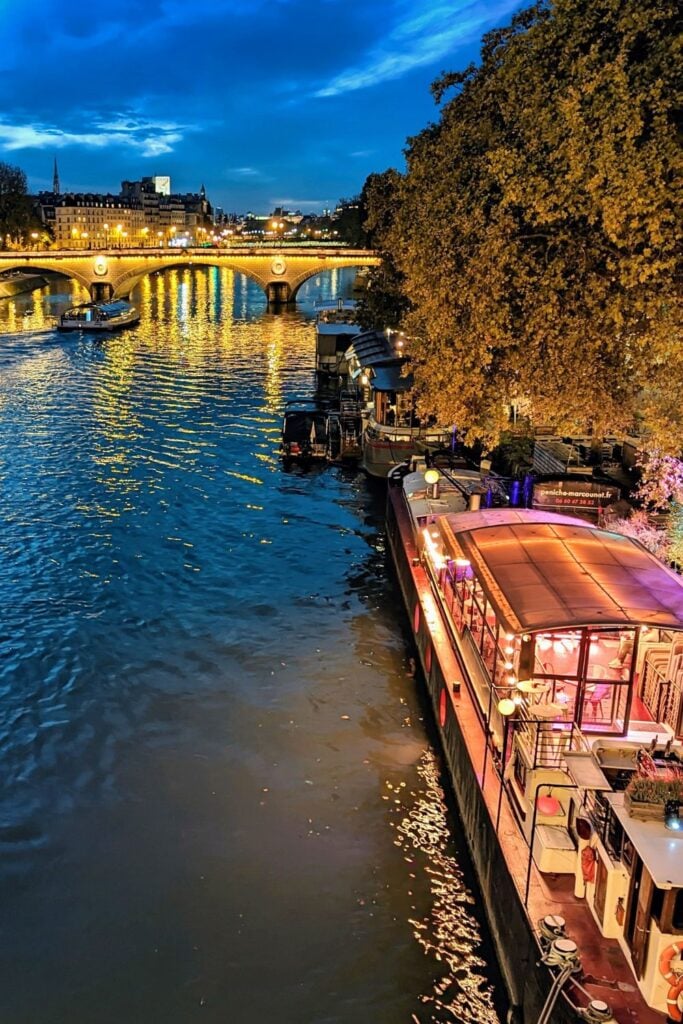 Illuminated restaurant boats on Seine River at night with bridge lights reflecting in water and historic buildings along riverbank