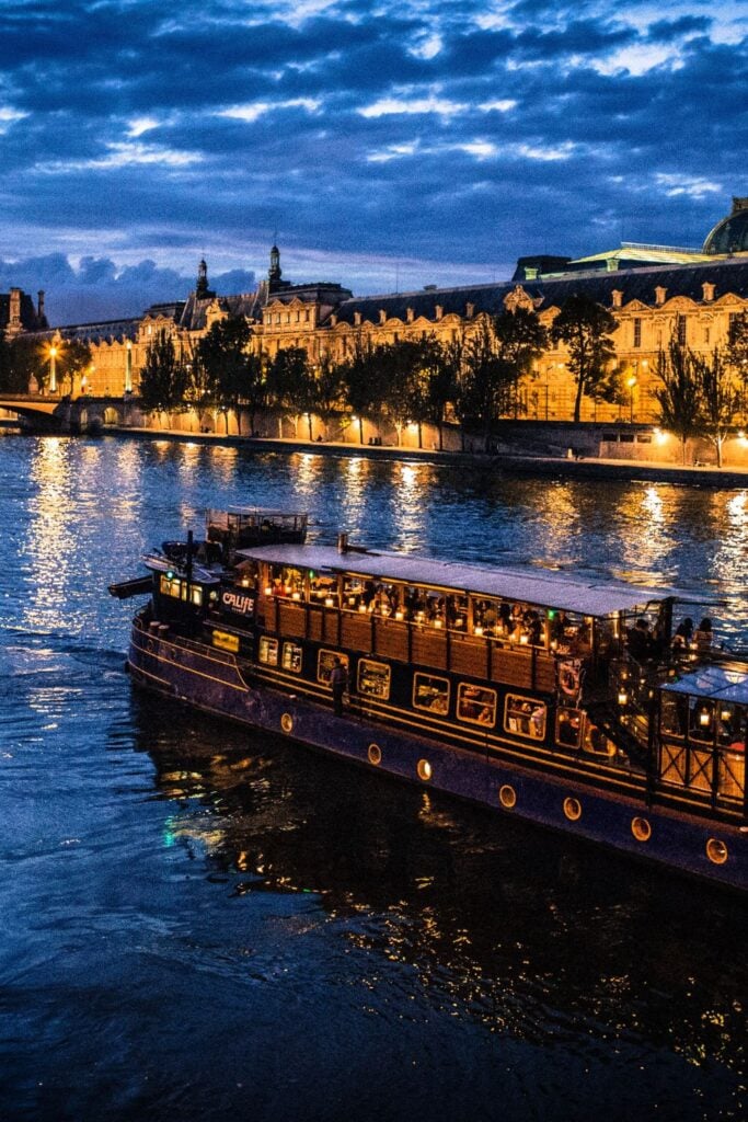 Illuminated dinner cruise boat on Seine River at night with historic buildings and their golden reflections on water