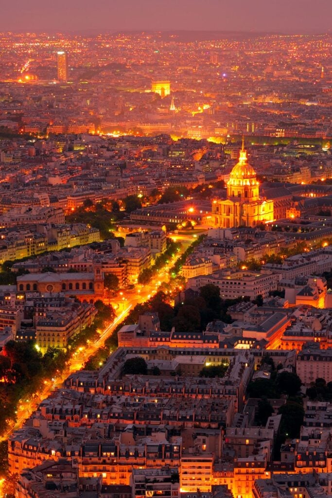 Aerial night view of Paris with golden lights illuminating the cityscape, featuring Les Invalides dome and Arc de Triomphe