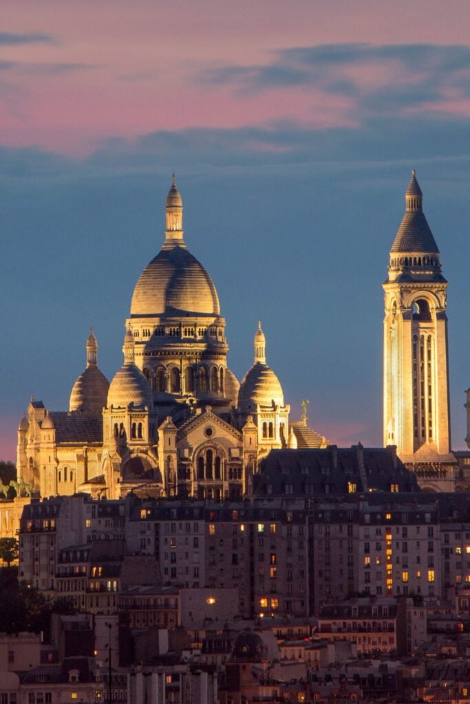 Sacré-Cœur Basilica illuminated at dusk with golden light against blue sky and Parisian apartment buildings with lights in windows below