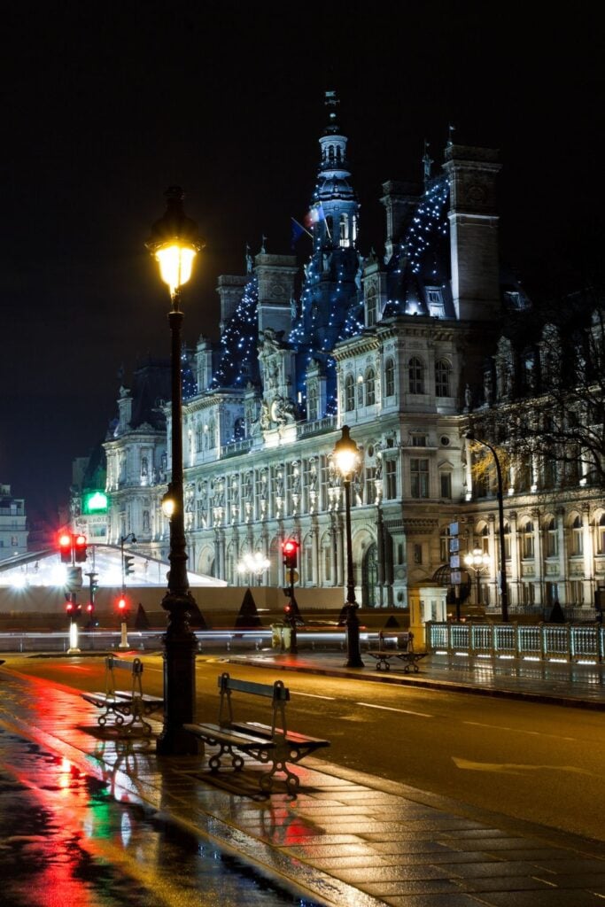 Hôtel de Ville Paris illuminated at night with blue lights and Christmas decorations along a rain-slicked street with colorful reflections