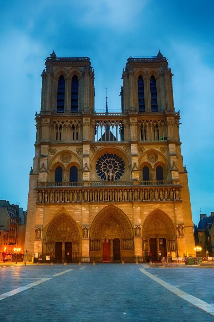 Notre Dame Cathedral façade illuminated in warm golden light against deep blue twilight sky with empty plaza in foreground