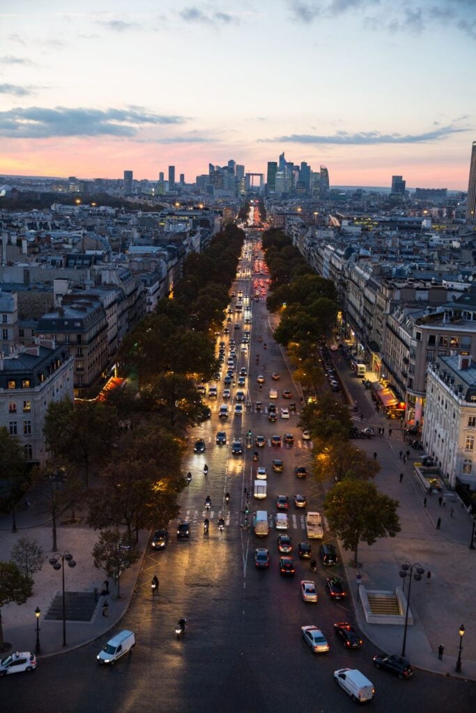 Aerial view of Champs-Élysées at dusk with streaming car lights leading toward La Défense district with pink sunset sky