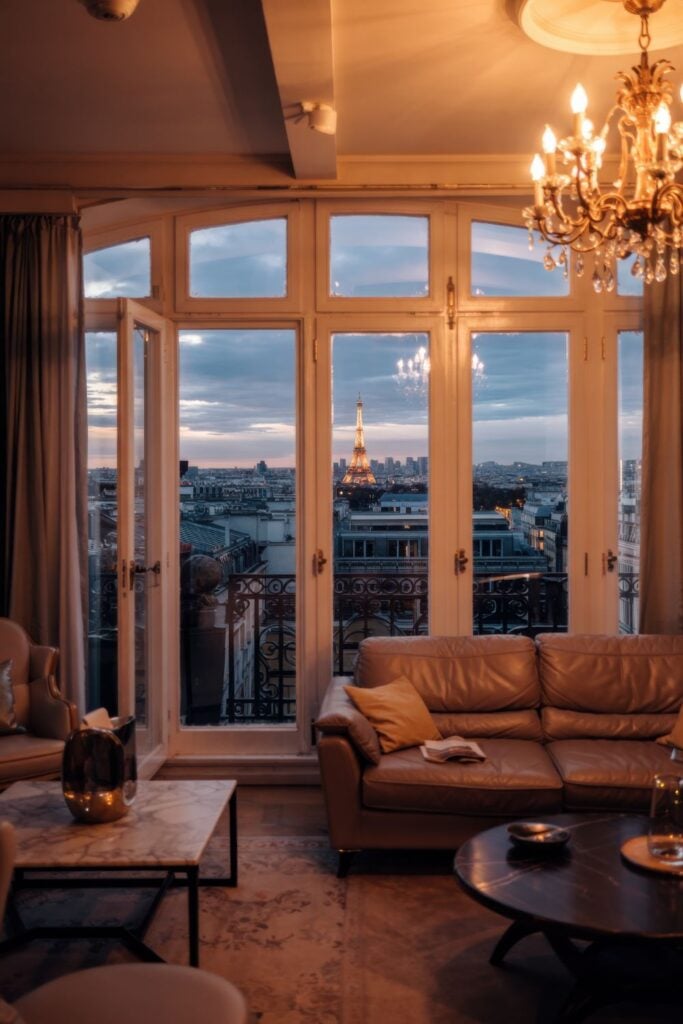 Luxury Paris apartment interior with French doors framing twilight view of illuminated Eiffel Tower with chandelier hanging in foreground