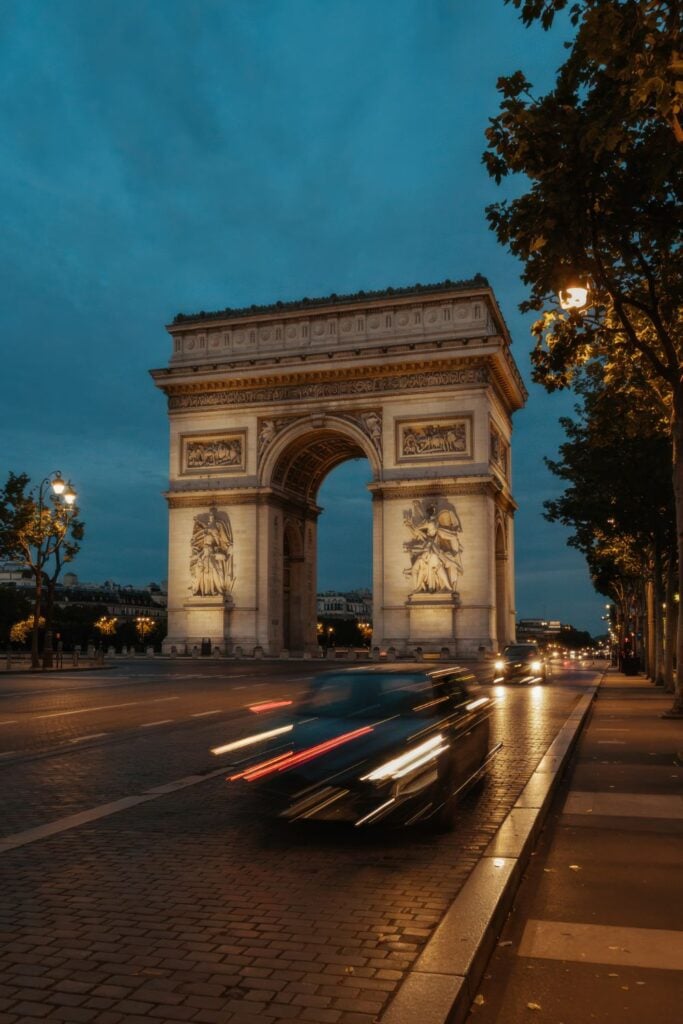 Arc de Triomphe illuminated at blue hour with motion-blurred car lights streaking past and tree-lined Champs-Élysées in foreground