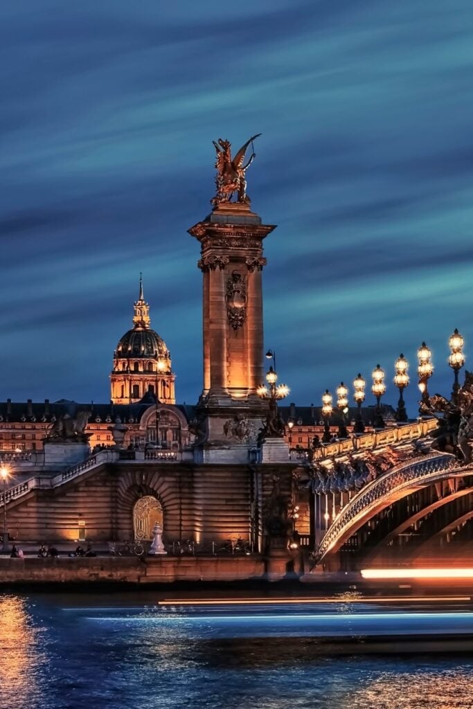 Pont Alexandre III bridge at night with illuminated golden statues and Les Invalides dome glowing in background against deep blue sky