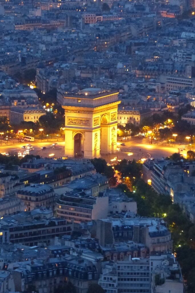 Aerial view of illuminated Arc de Triomphe at night with glowing streets radiating outward and city lights spread across Paris
