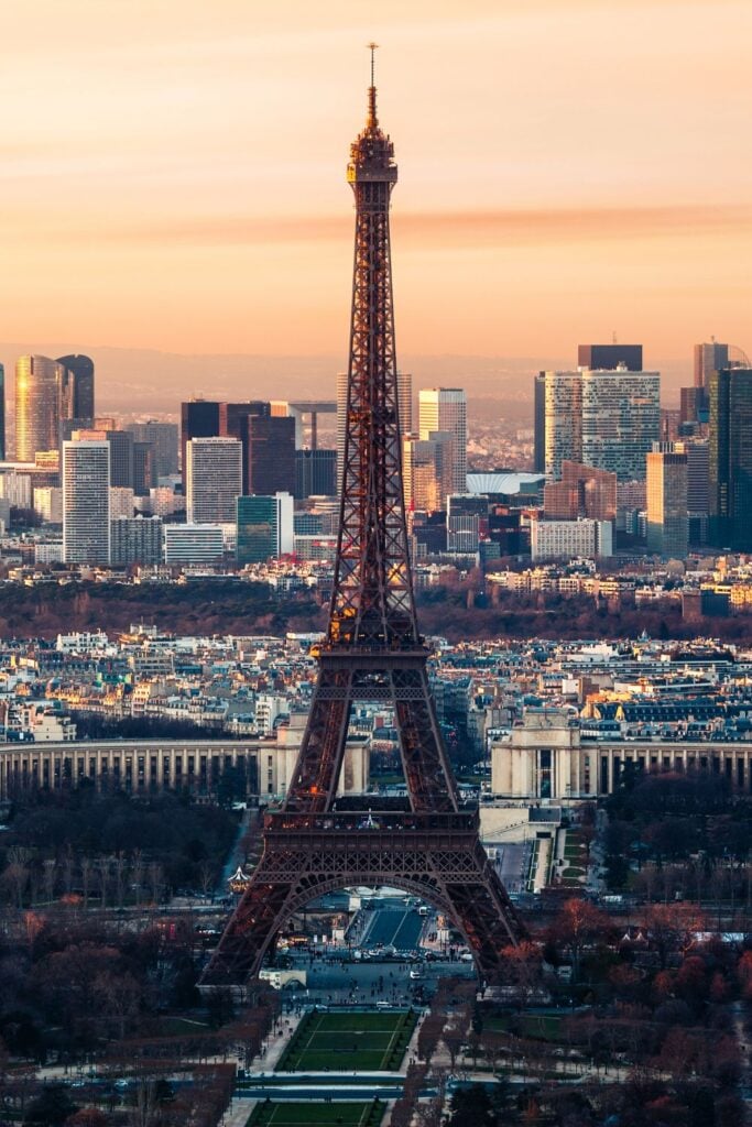 Eiffel Tower at sunset with La Défense business district and Parisian skyline in background against orange-peach sky
