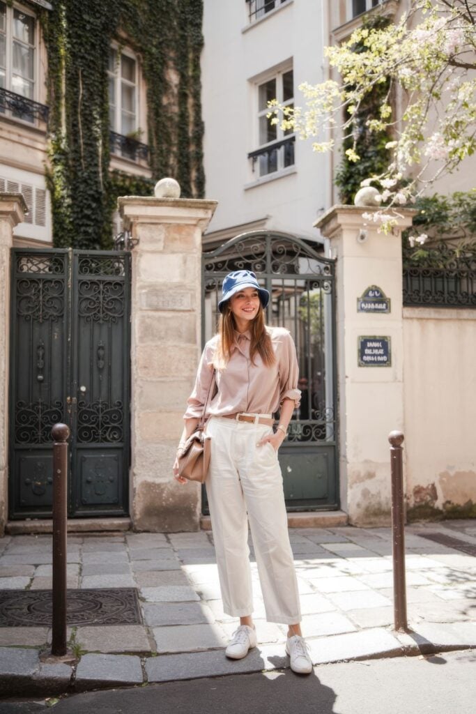 Woman wearing blush silk shirt, cream trousers, blue bucket hat and white sneakers in front of ornate Parisian courtyard entrance