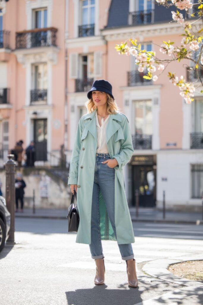 Woman in mint green trench coat with navy bucket hat, white shirt and blue jeans against pink Parisian buildings