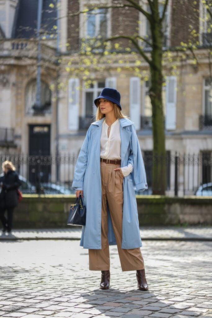 Woman wearing powder blue trench coat with white shirt and camel trousers on cobblestone street with budding trees in background