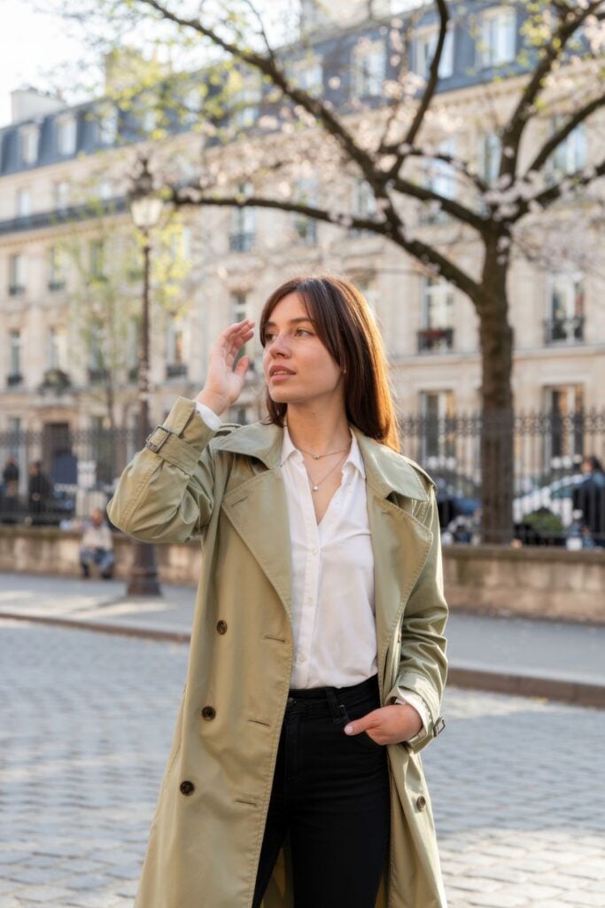 Woman in beige trench coat, white shirt and black pants standing on cobblestone street with cherry blossoms in background