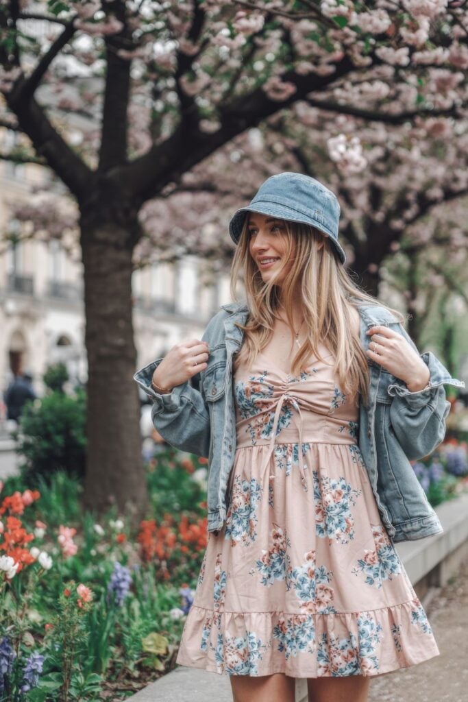Woman wearing floral pink dress with denim jacket and blue bucket hat standing beside colorful flower garden with cherry blossom trees overhead