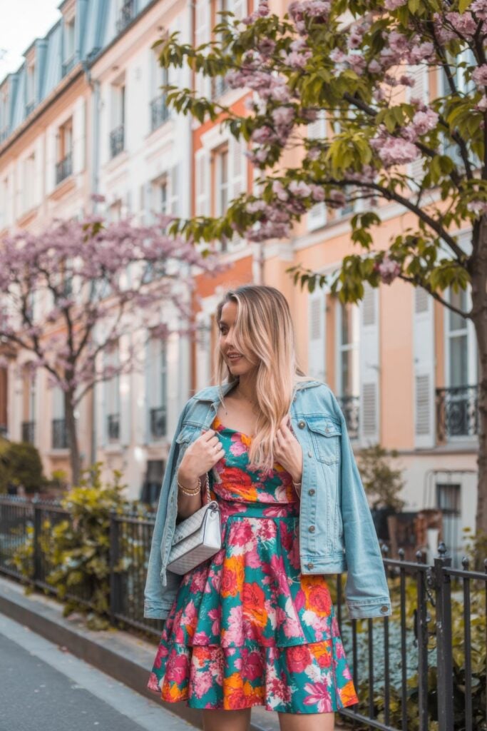 Woman wearing vibrant floral teal dress with light denim jacket standing on Parisian street with pink buildings and cherry blossoms