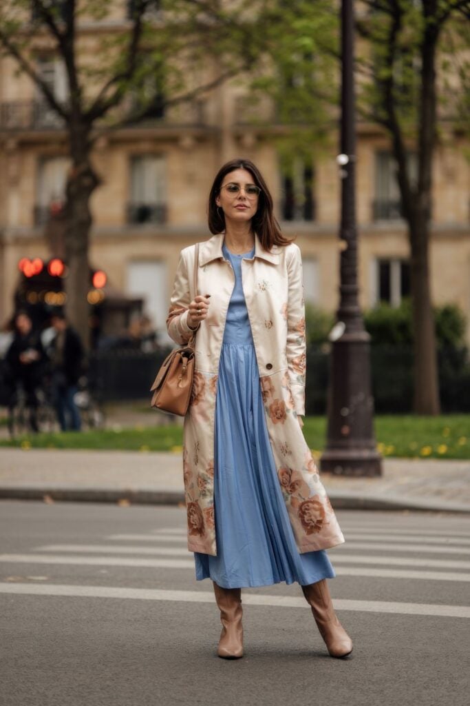 Woman wearing cream floral trench coat with powder blue dress crossing Parisian street with budding trees in background