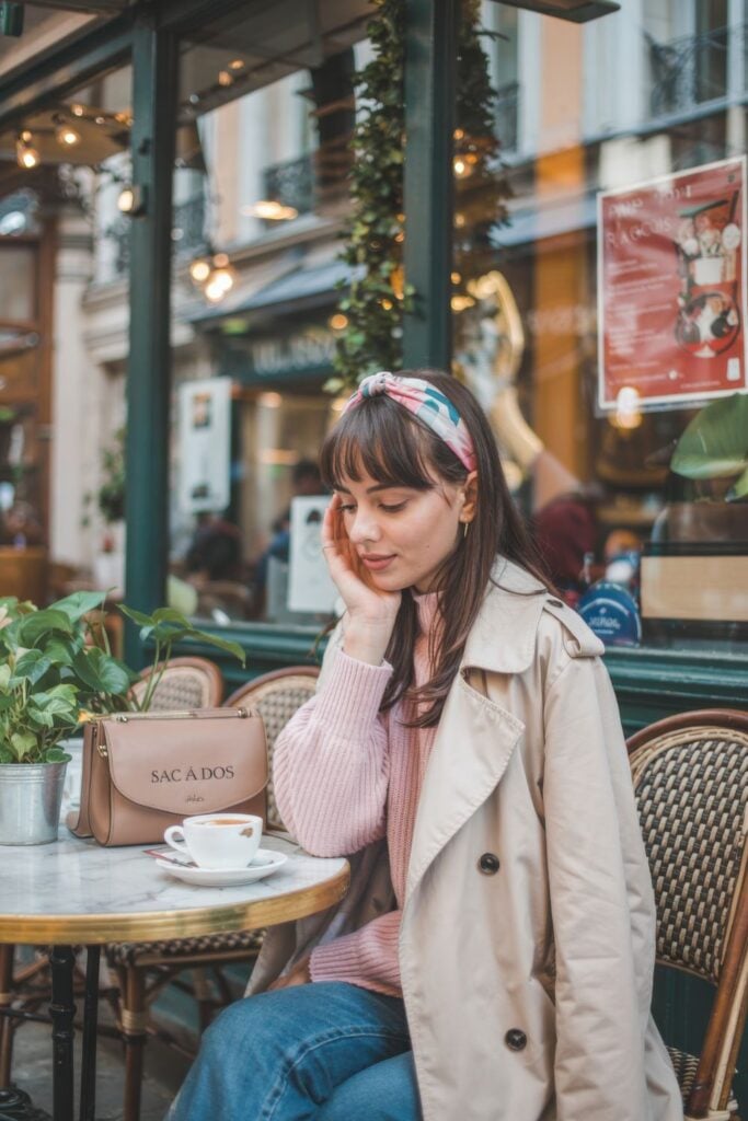 Woman in beige trench coat and pink sweater with printed headband sitting at outdoor Parisian café terrace with espresso