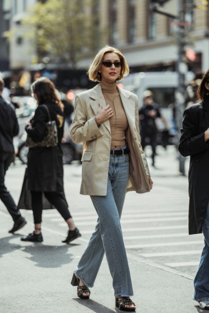 Woman in beige blazer and camel turtleneck walking across a busy Paris street in wide-leg jeans and sunglasses