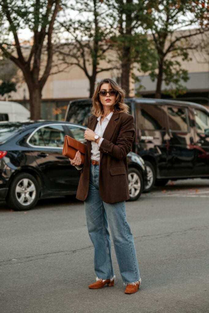 Woman in brown blazer, white shirt, and jeans holding a clutch while walking through a Paris neighborhood