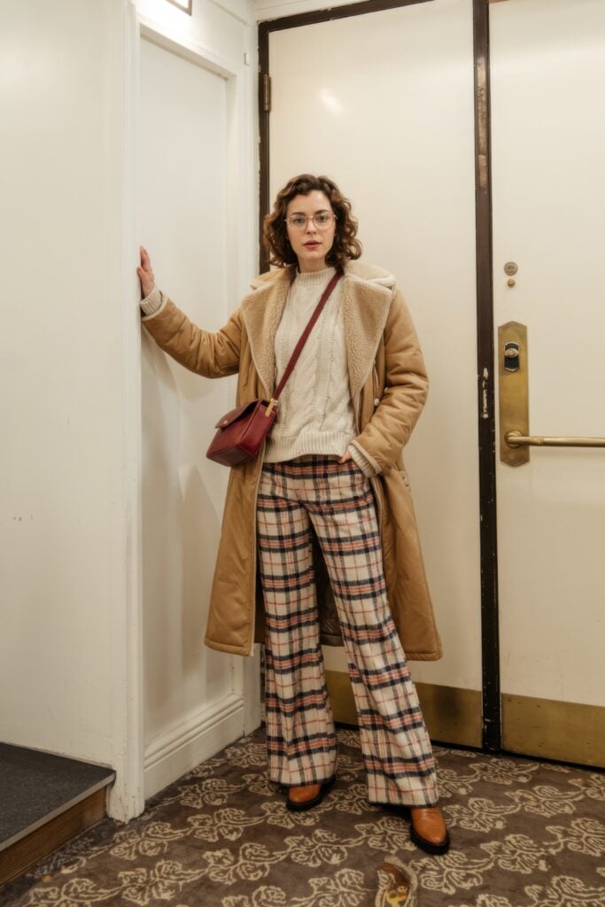 Woman in shearling coat, cream sweater, and plaid pants posing indoors with a relaxed, retro vibe