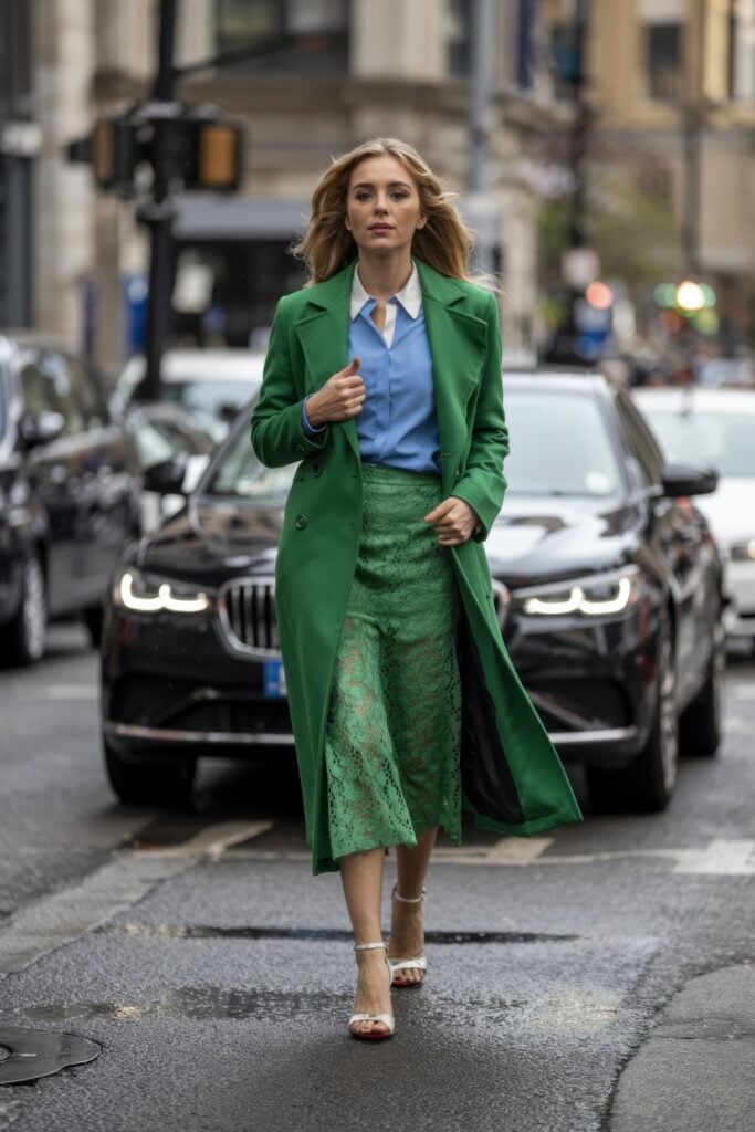 Woman in emerald green lace skirt and coat paired with a blue blouse crossing a wet city street in heels