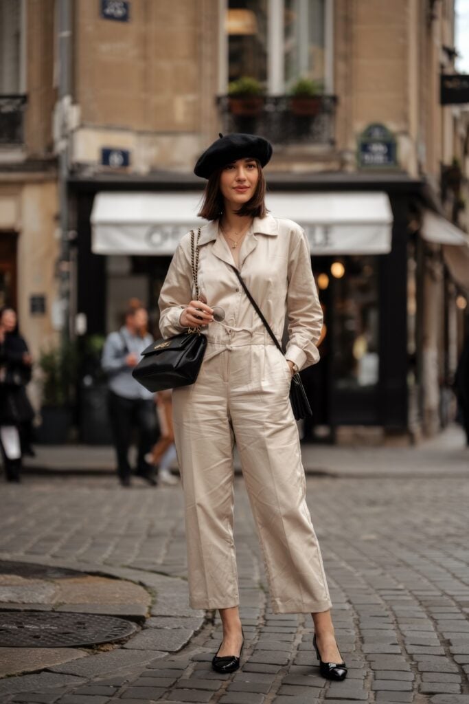 Woman in cream jumpsuit and black beret standing on a cobbled street in Paris with a black handbag