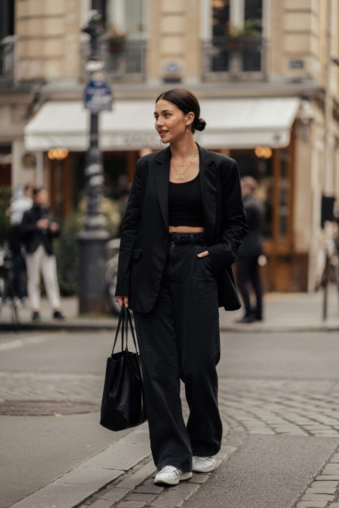 Woman in an all-black outfit with a black blazer, crop top, wide-leg jeans, and white sneakers, walking through a Paris intersection.