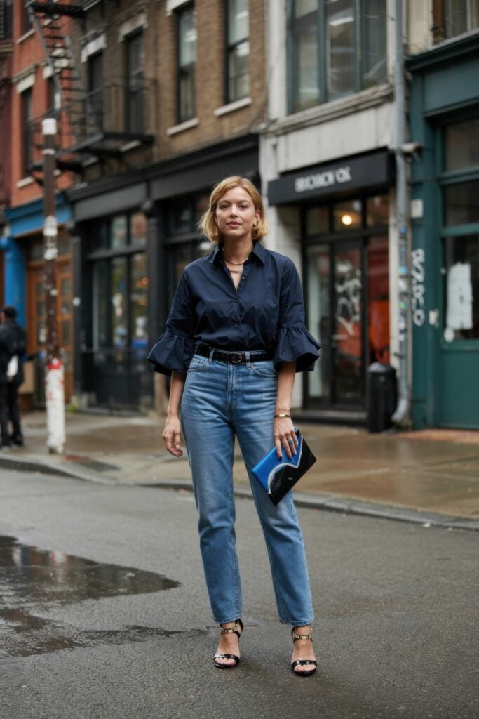 Woman in navy ruffle-sleeve blouse and high-waisted jeans holding a clutch on a Paris side street