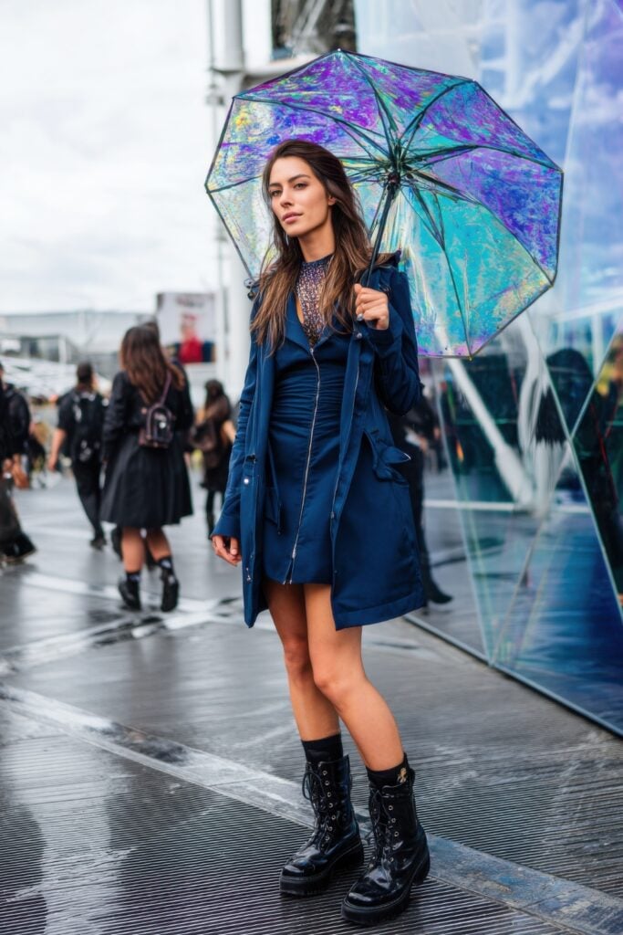 Woman in a navy trench and ruched dress, combat boots, and a holographic umbrella walking across a rainy fashion district in Paris.