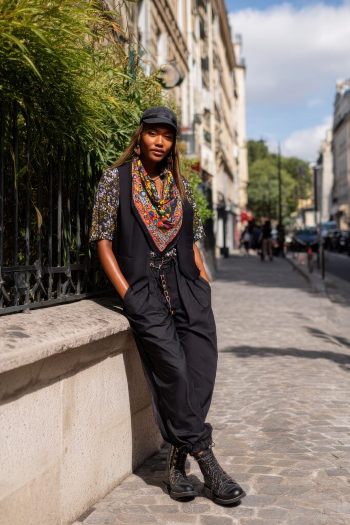Woman leaning on a stone wall in black joggers, a colorful scarf, floral shirt, vest, and lace-up boots on a Paris side street.
