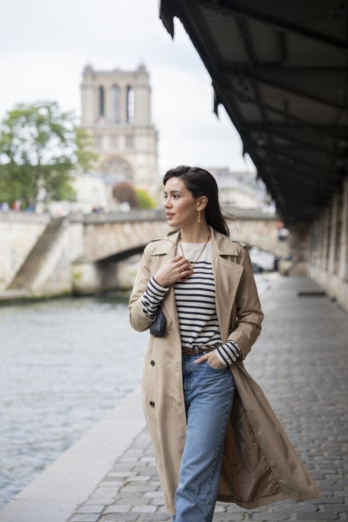 Woman in a tan trench coat, striped top, and jeans walking along the Seine near Notre-Dame Cathedral in Paris.