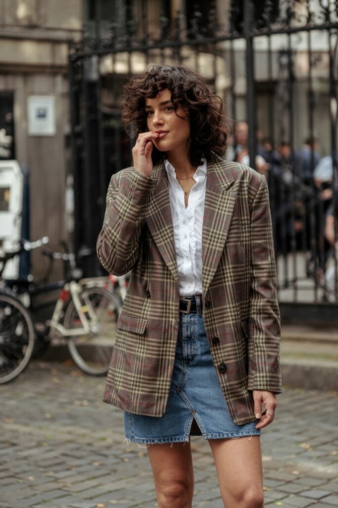 Woman in oversized plaid blazer and raw-hem denim skirt with white shirt posing on a cobbled street in Paris