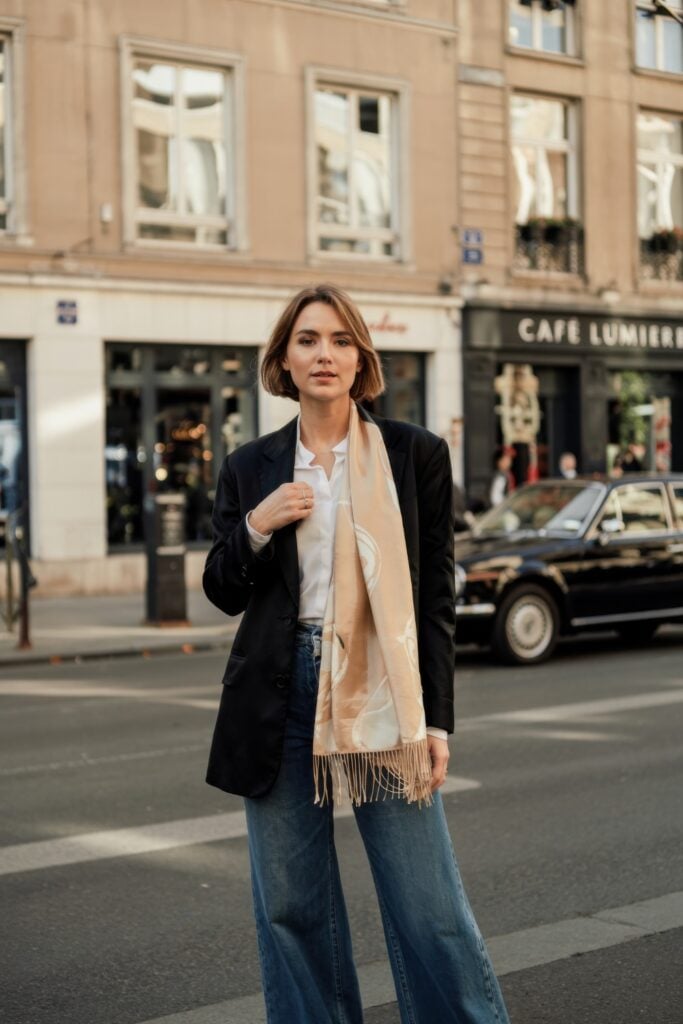 Woman in wide-leg jeans, black blazer, and neutral silk scarf standing in front of a Parisian café