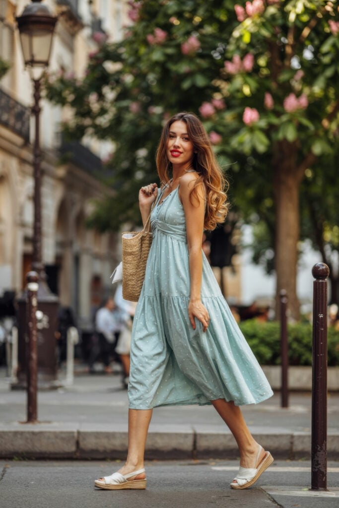 Woman wearing light blue polka-dot sundress with tiered skirt and straw bag walking on Parisian street