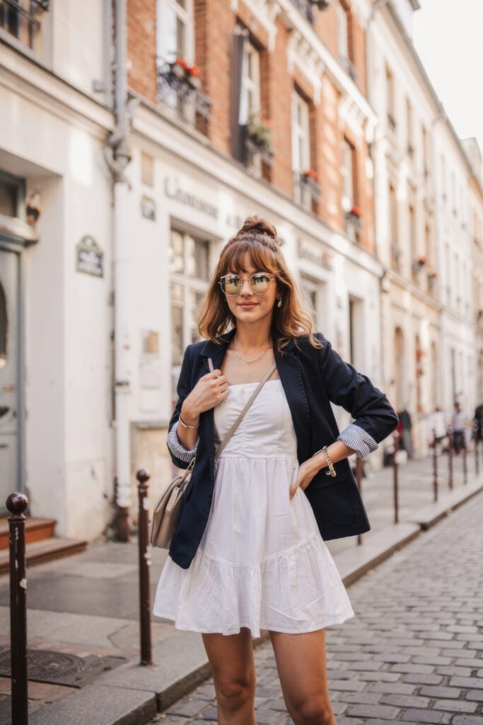 Woman wearing white sundress with navy blazer and eyeglasses on cobblestone Parisian street