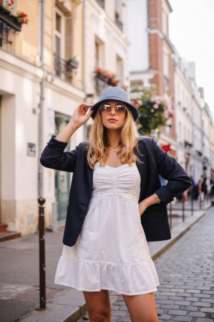 Woman in white dress with navy blazer and matching bucket hat on Paris cobblestone street