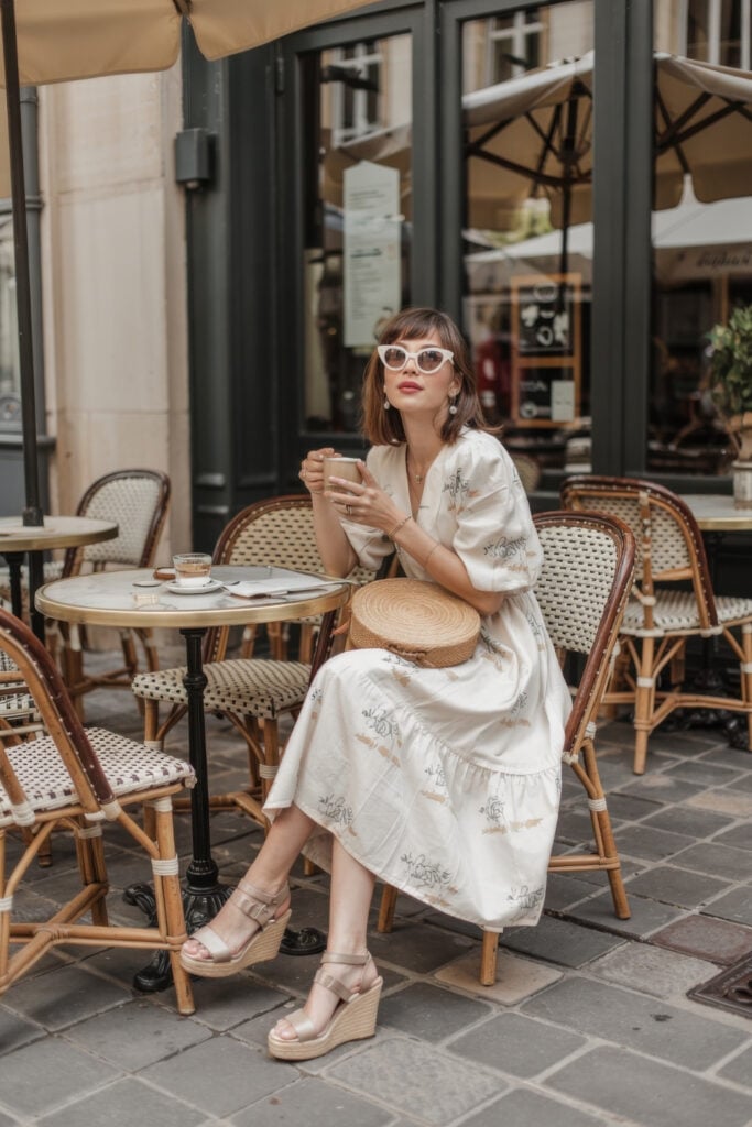 Woman in a Paris summer outfit with patterned white midi dress sitting at outdoor Parisian café with straw hat and coffee