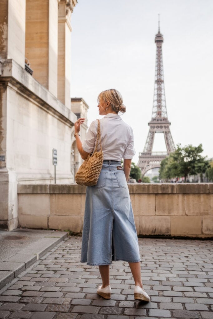 Back view of woman in white shirt and denim midi skirt with straw bag looking at Eiffel Tower