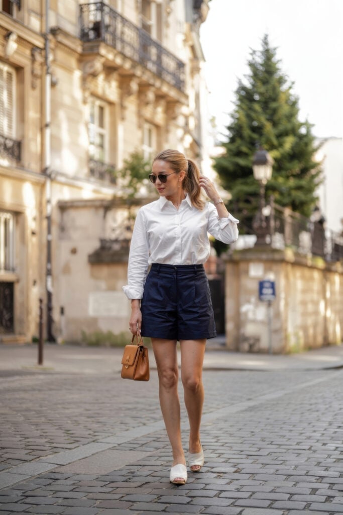 Woman in white button-down shirt with navy shorts and tan handbag walking on Paris street