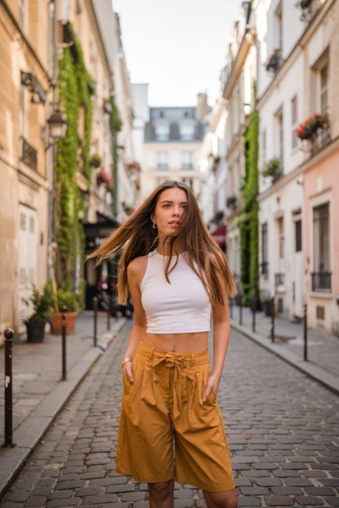 Woman wearing white crop top and mustard culottes walking down narrow Parisian street with ivy-covered buildings