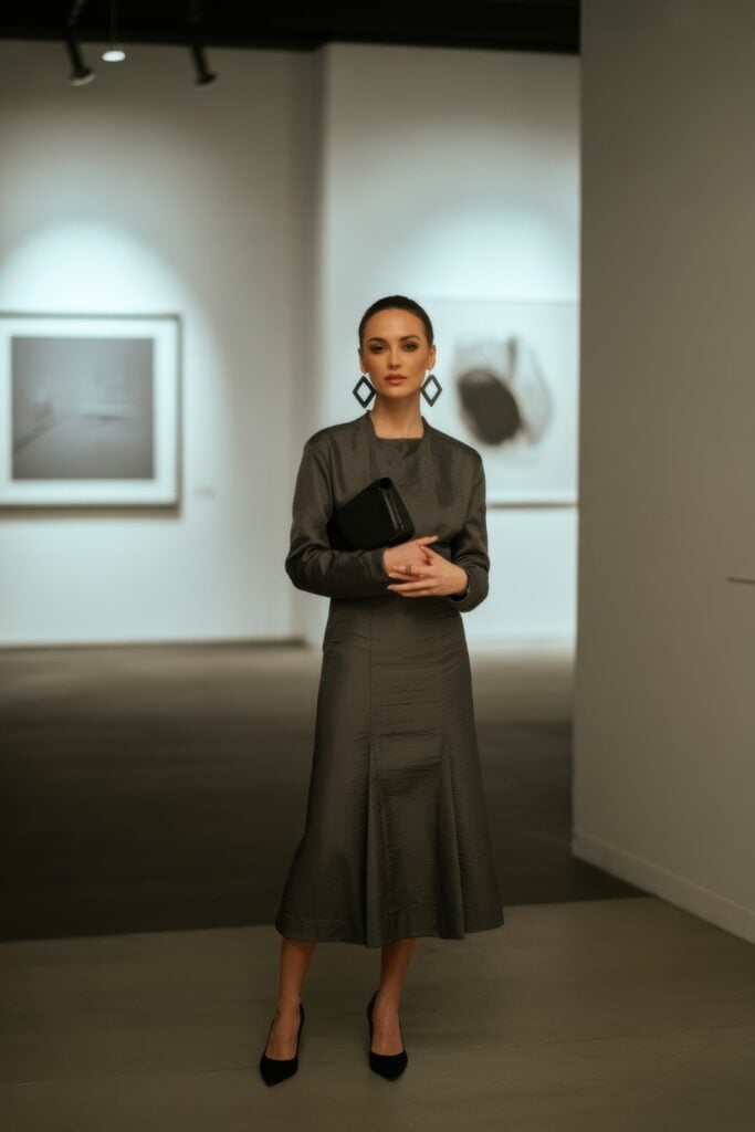Woman wearing dark olive green midi dress with structured silhouette in art gallery holding black clutch