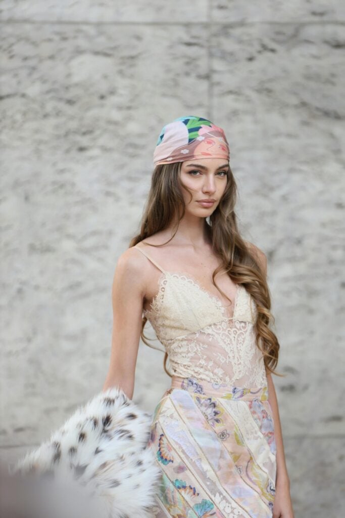 Woman in lace camisole with colorful patterned skirt, headscarf and spotted fur clutch against stone wall backdrop