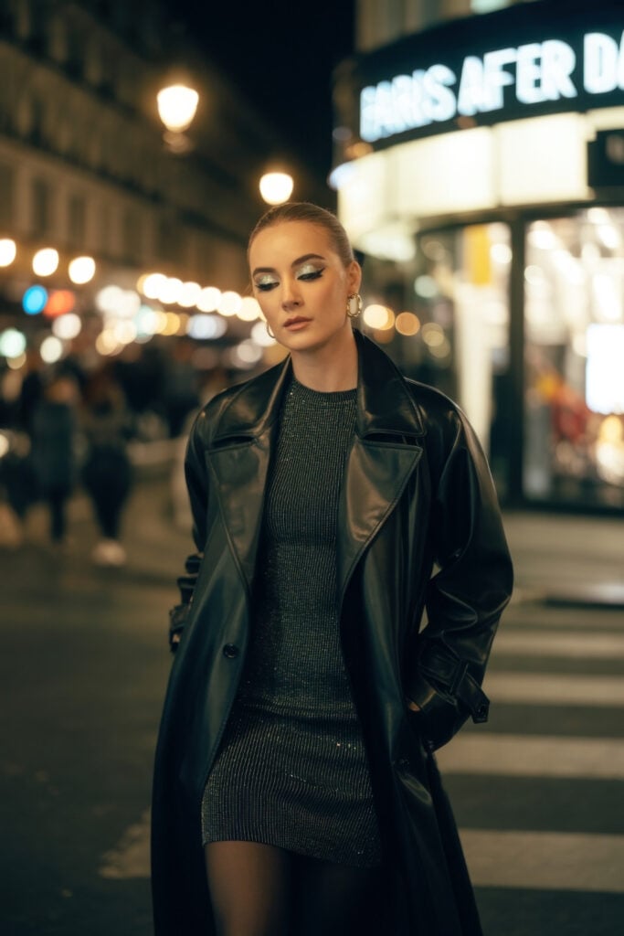 Woman in black leather trench coat over metallic dress with slicked-back hair on nighttime Paris street with neon lights