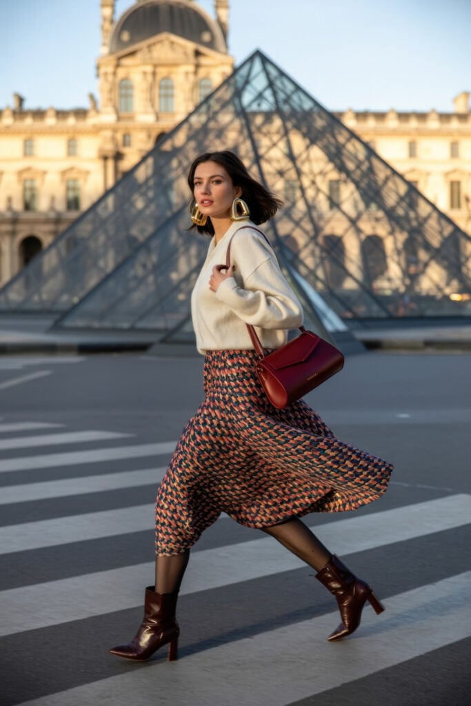 Woman in cream sweater and geometric print midi skirt with red handbag walking in front of Louvre pyramid