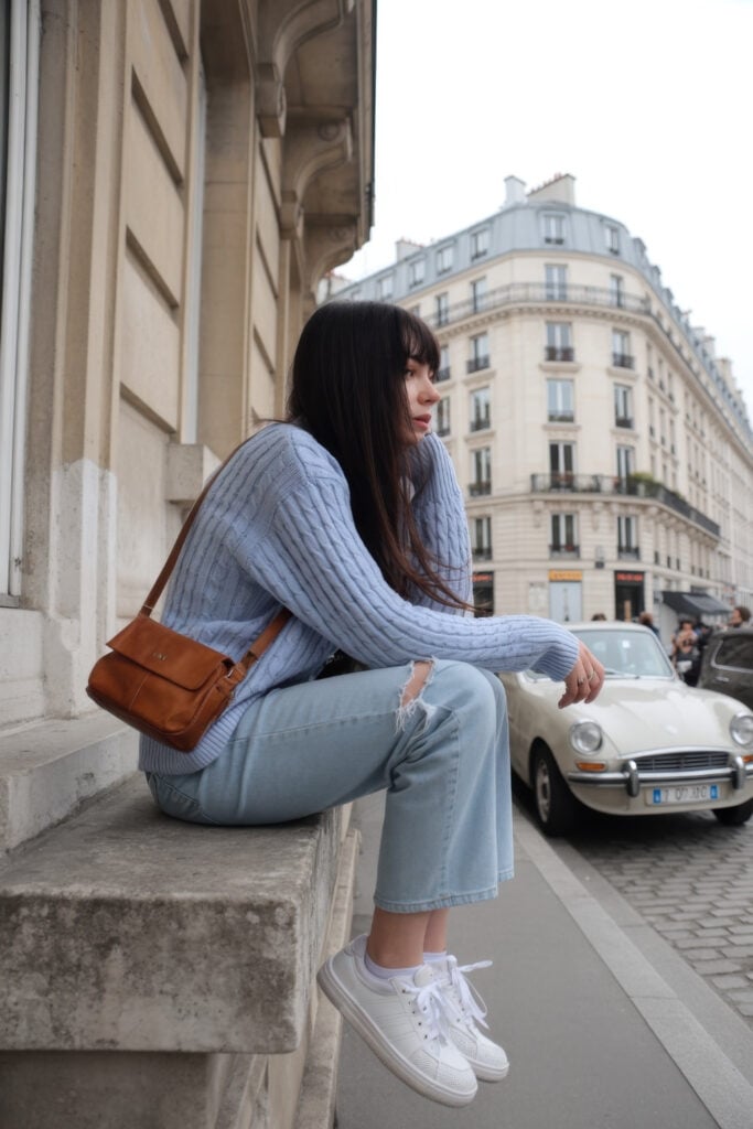 Woman in light blue cable-knit sweater and distressed jeans with brown crossbody bag sitting on Parisian street ledge with vintage car
