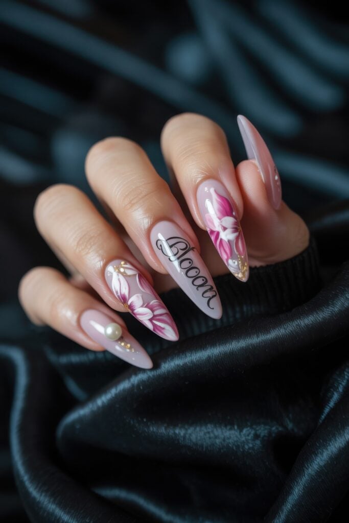Long almond-shaped nails with pink and white base, featuring "Amour" script lettering, pink floral nail art, and pearl accents on dark background.