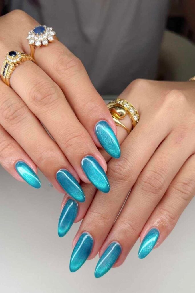 Long oval nails in shimmery turquoise blue polish styled with ornate gold rings featuring blue stones and diamonds against neutral background.