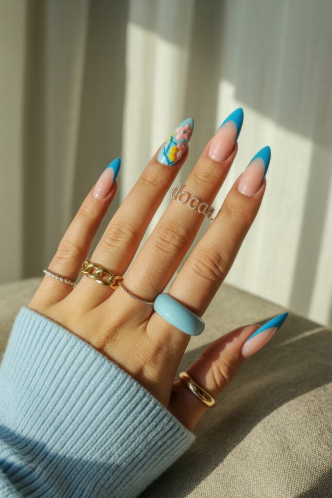 Almond-shaped nails, bright blue tips, and cartoon character nail art, styled with multiple gold and silver rings in natural lighting.