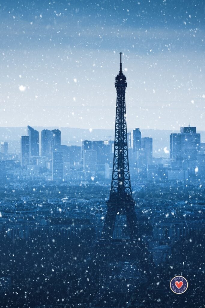 Eiffel Tower silhouetted against blue winter sky with heavy snowfall over Paris cityscape with La Défense buildings in background