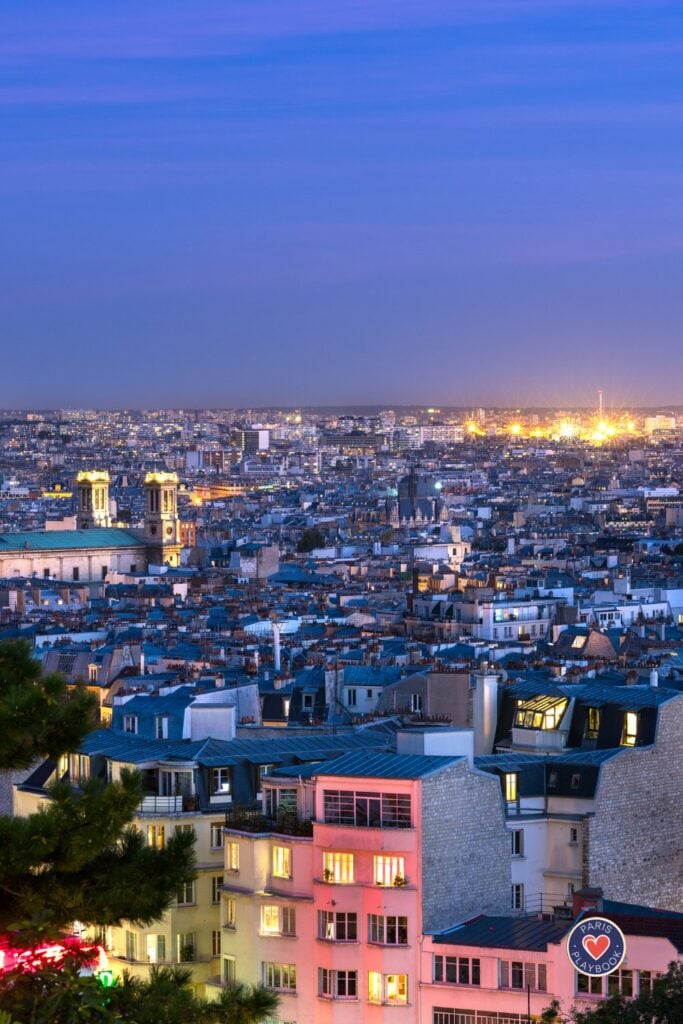 Aerial view of Paris rooftops at blue hour with illuminated windows creating a sea of lights across residential buildings with Notre Dame towers visible in distance against purple evening sky