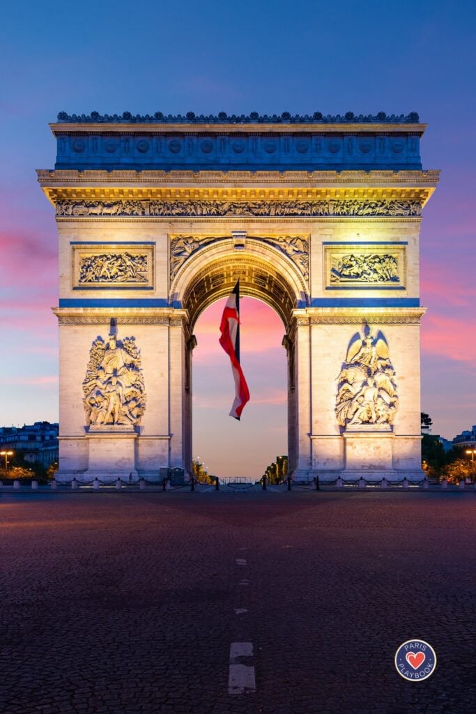 Arc de Triomphe illuminated at blue hour with French tricolor flag waving beneath the arch against dramatic pink and purple sunset sky on Place Charles de Gaulle